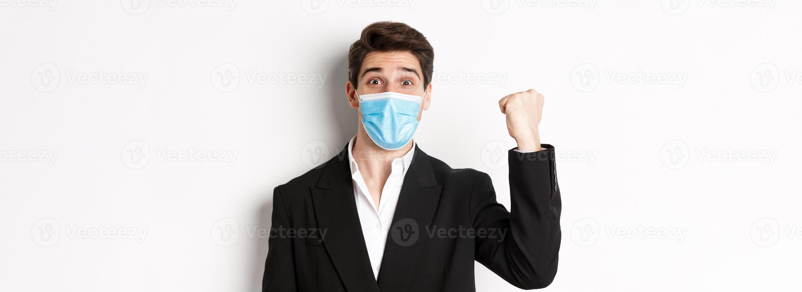 Concept of covid-19, business and social distancing. Close-up of cheerful businessman in medical mask and suit, rejoicing, achieving goal and celebrating, standing over white background photo