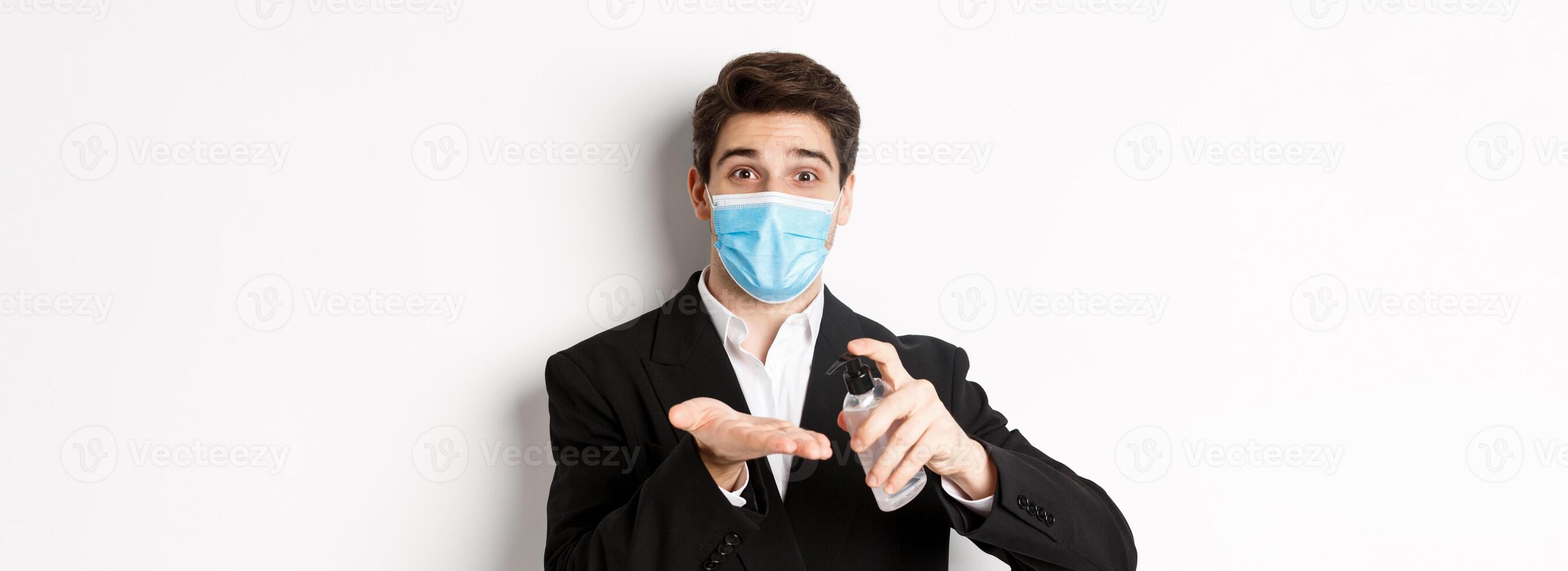 Concept of covid-19, business and social distancing. Image of handsome businessman in trendy suit and medical mask, cleaning hands with hand sanitizer, standing over white background photo