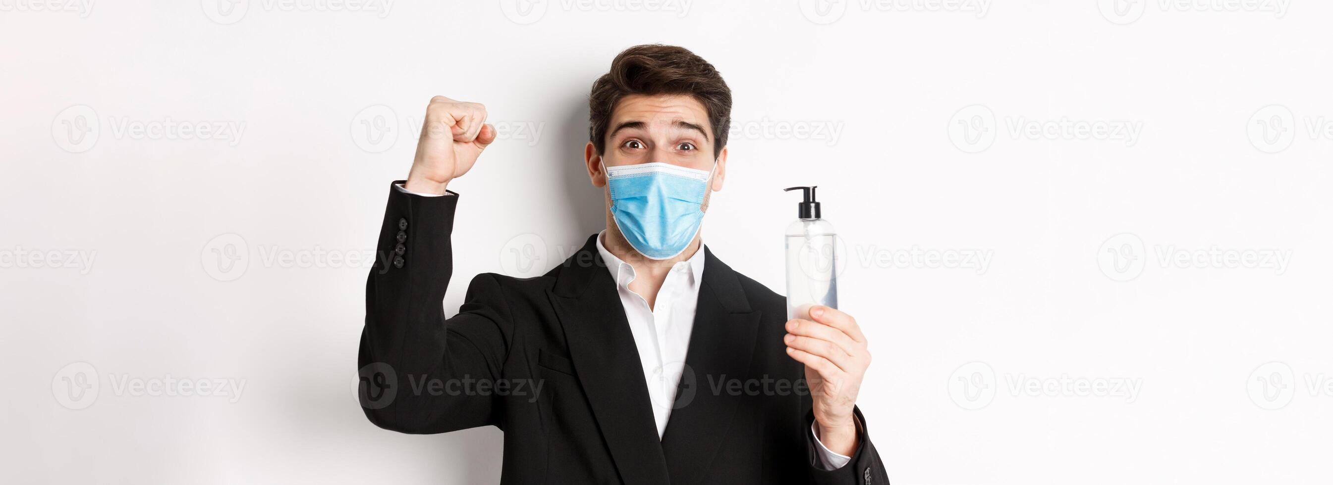 Concept of covid-19, business and social distancing. Close-up of happy man in trendy suit and medical mask, cheering and raising hand up, showing hand sanitizer, white background photo