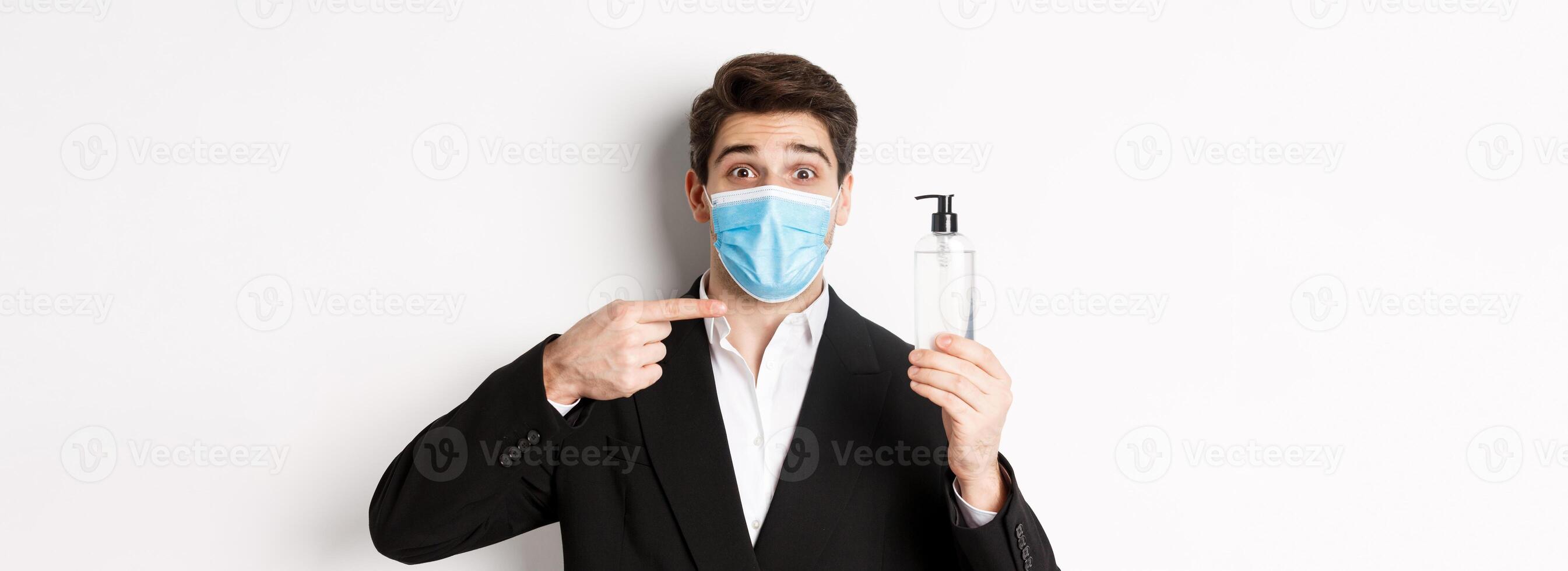 Concept of covid-19, business and social distancing. Close-up of handsome man in trendy suit and medical mask, recommending hand sanitizer, standing against white background photo
