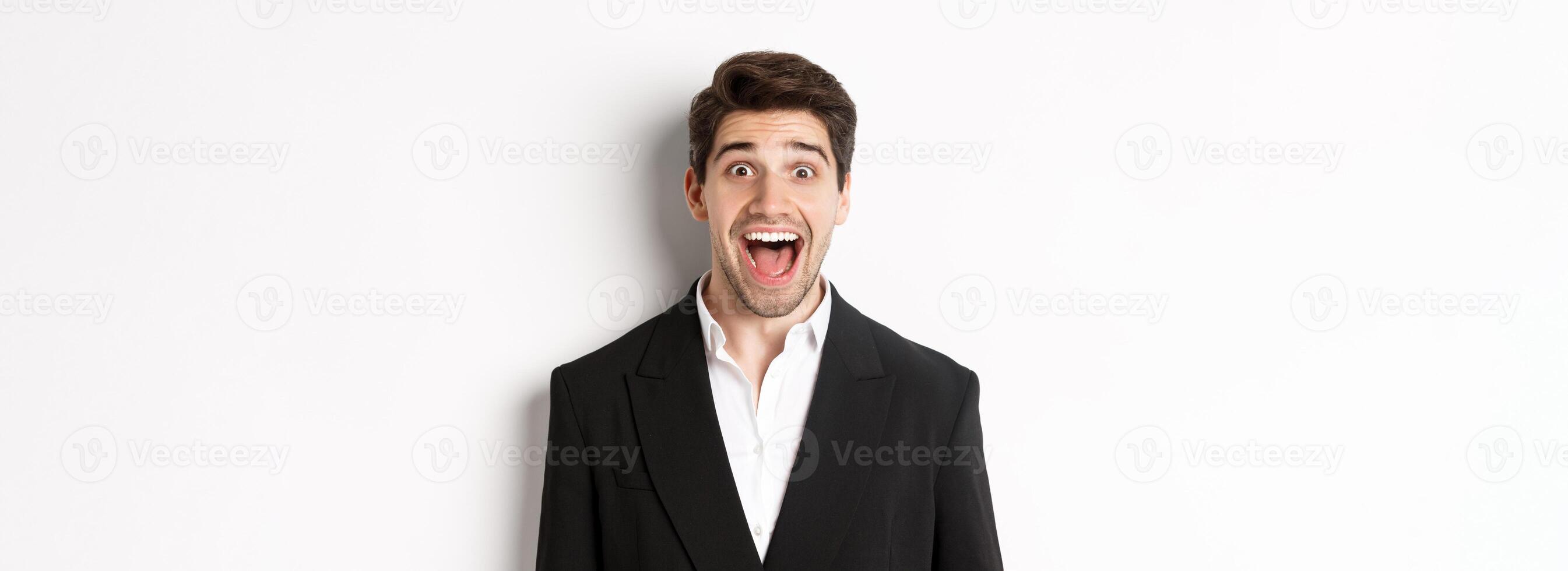 Close-up of attractive man in black suit, smiling amazed and looking at advertisement, standing over white background photo
