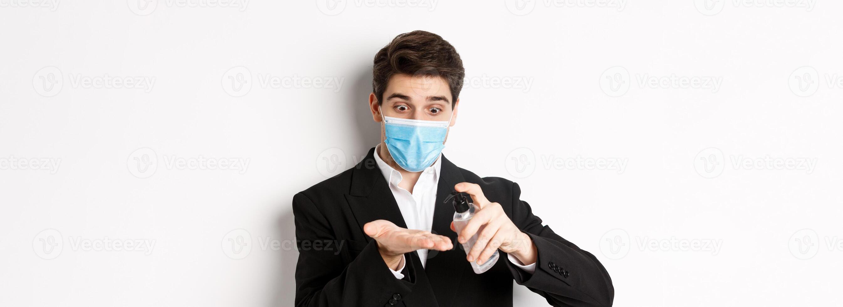 Concept of covid-19, business and social distancing. Image of handsome businessman in trendy suit and medical mask, cleaning hands with hand sanitizer, standing over white background photo