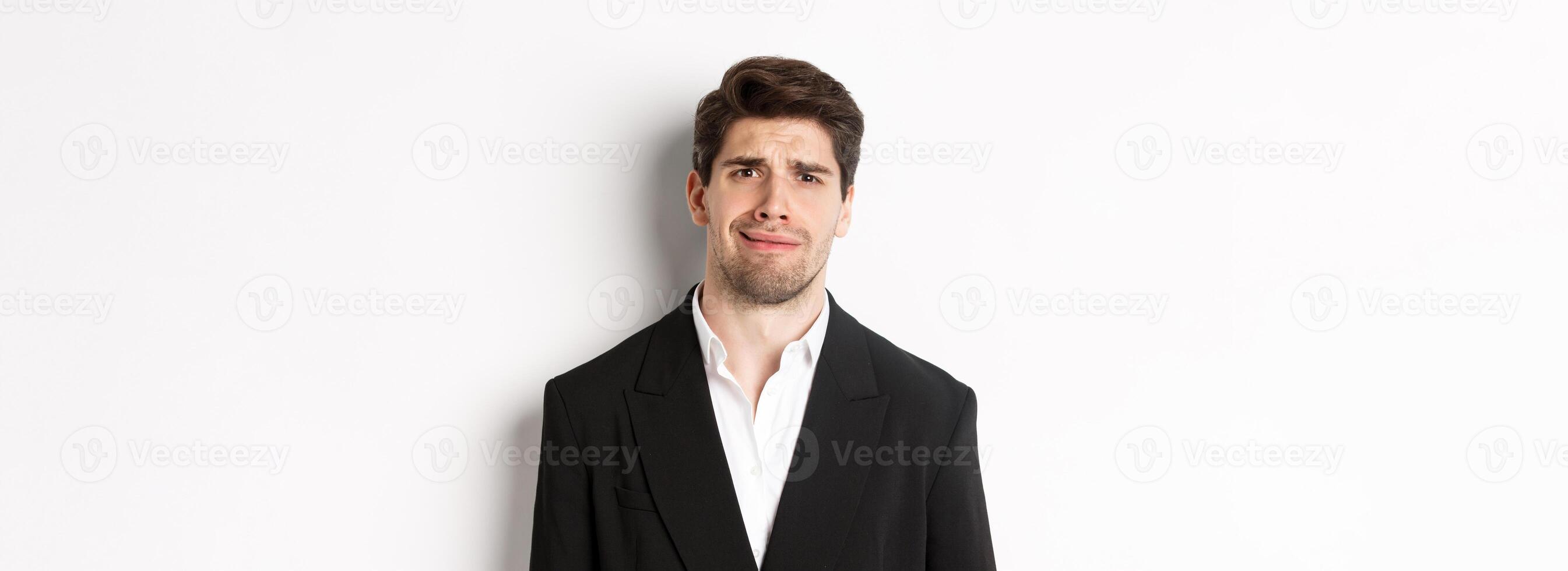 Close-up of confused handsome man in suit, grimacing and looking perplexed, wearing trendy suit, standing against white background photo