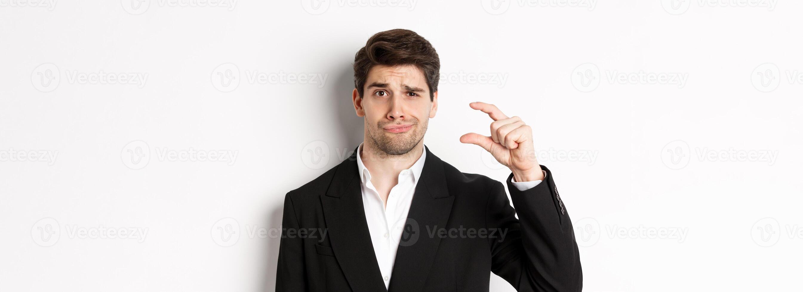 Close-up of handsome businessman in trendy suit, showing something small and looking disappointed, standing against white background photo