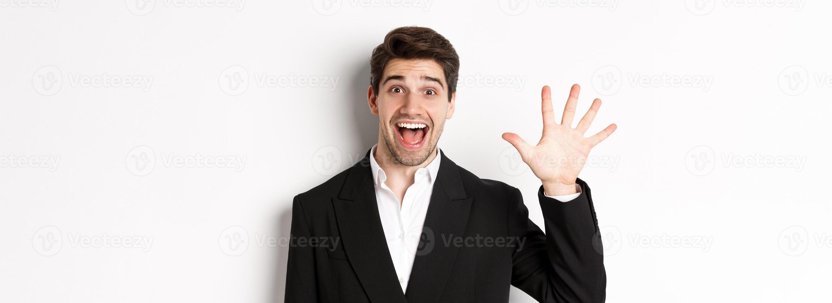 Close-up of handsome businessman in black suit, smiling amazed, showing number five, standing over white background photo