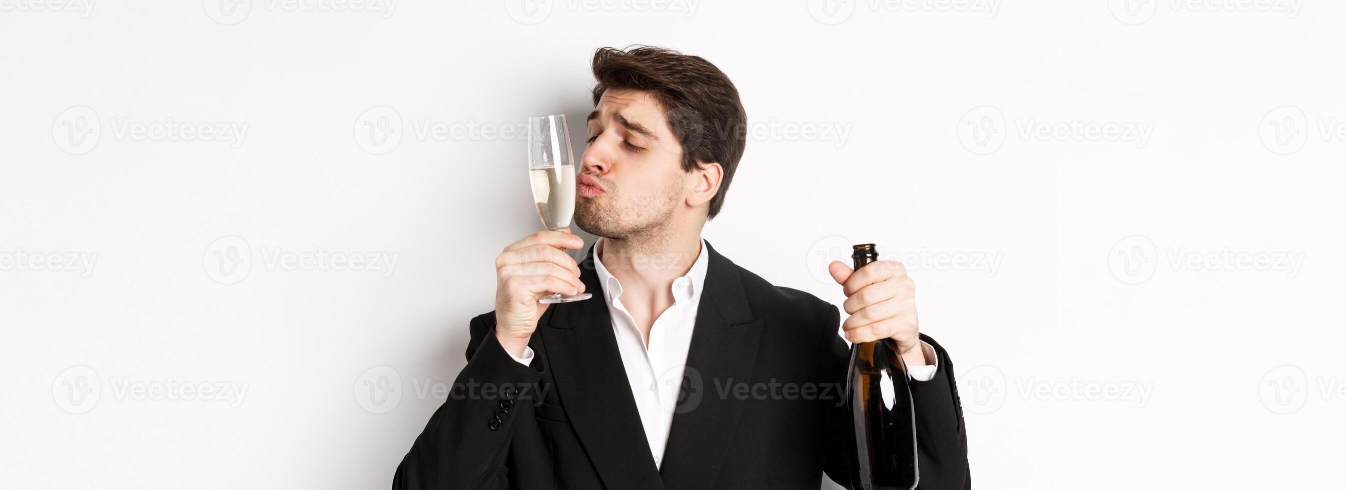 Close-up of handsome man in suit, kissing glass with champagne, getting drunk on a party, standing against white background photo