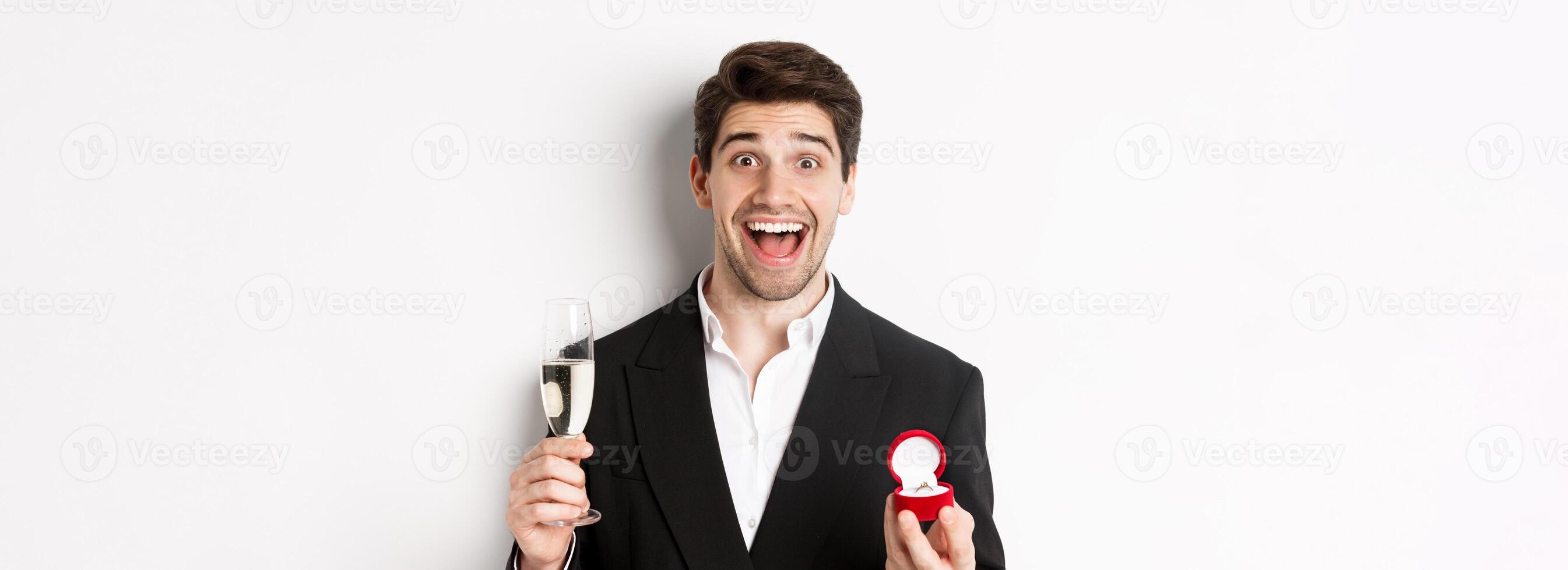 Close-up of handsome man in suit, making a proposal, giving engagement ring and raising glass of champagne, standing against white background photo
