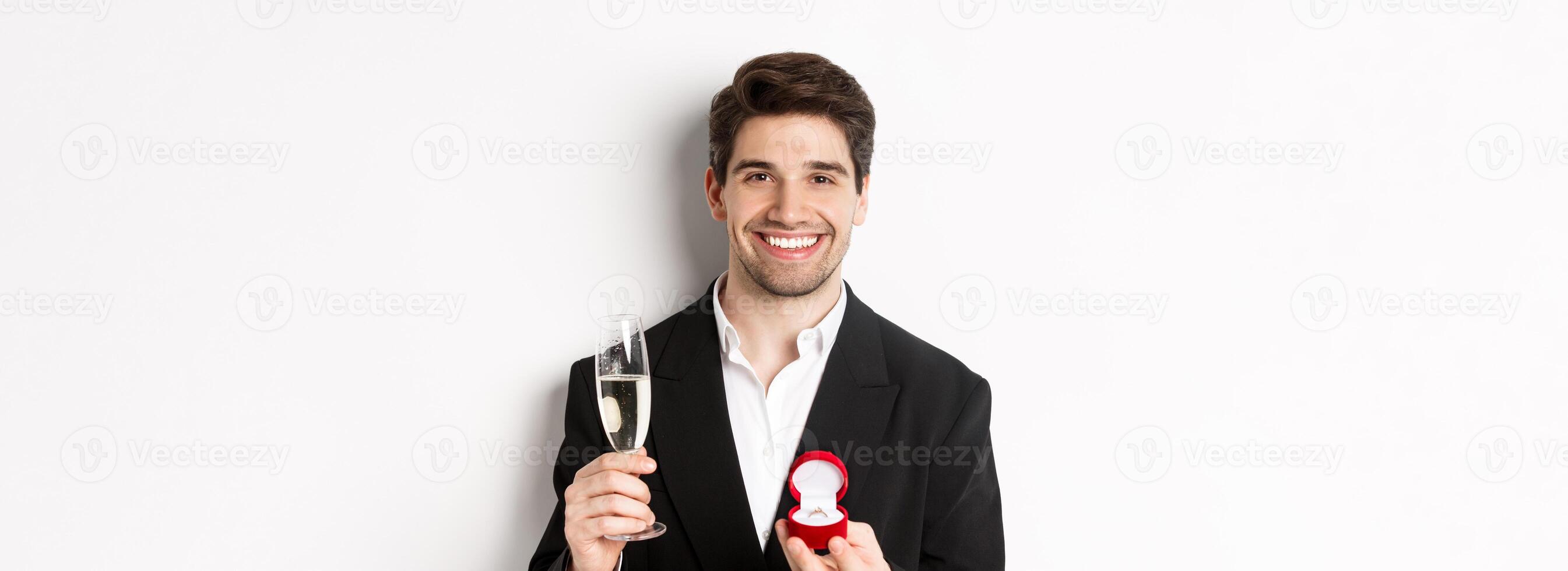 Close-up of handsome man in suit, making a proposal, giving engagement ring and raising glass of champagne, standing against white background photo