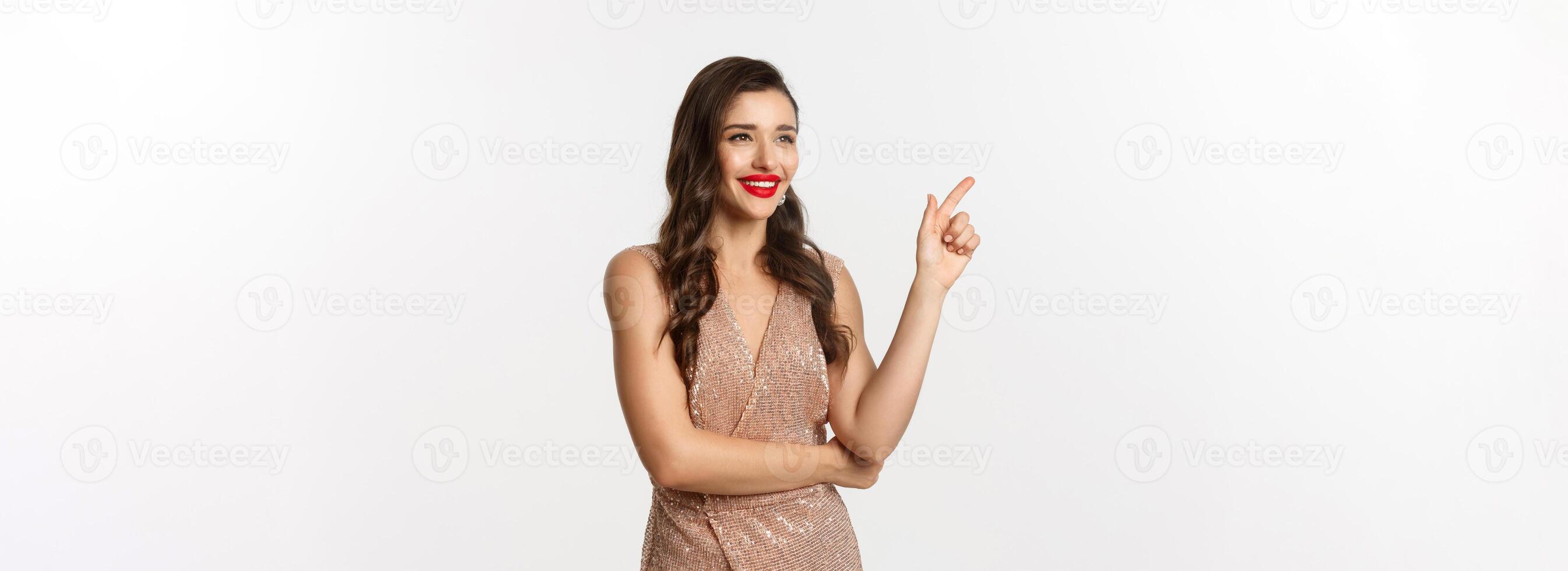 Christmas, holidays and celebration concept. Attractive woman wearing party dress and red lipstick, pointing and looking left, enjoing new year eve, white background photo