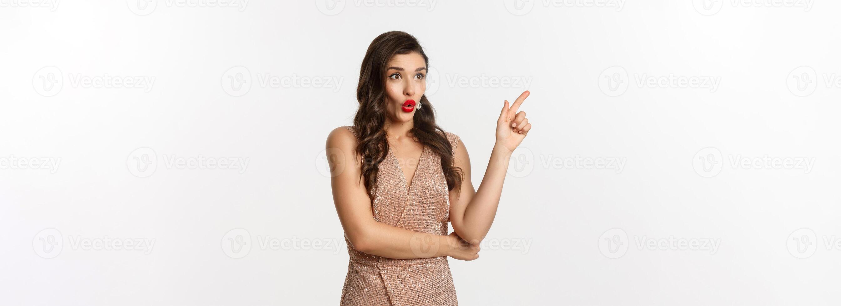 Christmas, holidays and celebration concept. Surprised girlfriend in evening dress, saying wow and pointing finger at promo offer, starring at logo, standing over white background photo
