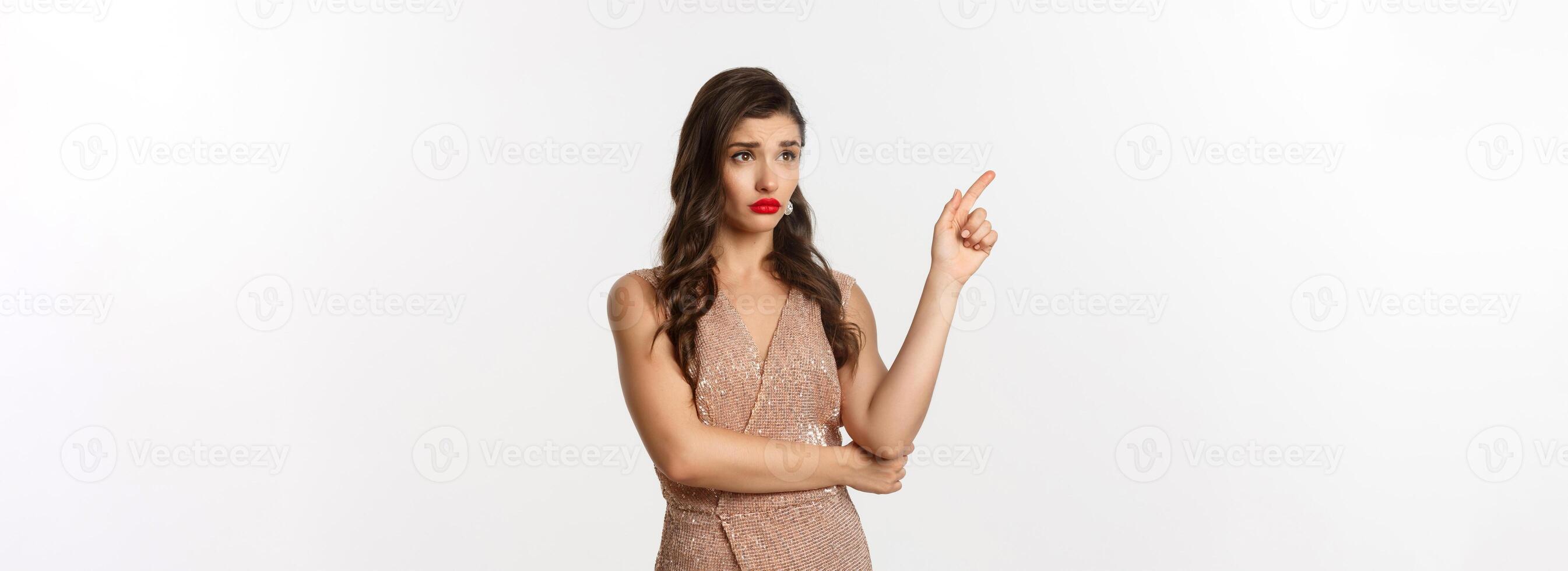 Christmas, holidays and celebration concept. Skeptical and displeased woman in stylish party dress, pointing finger left and starring at banner unamused, white background photo