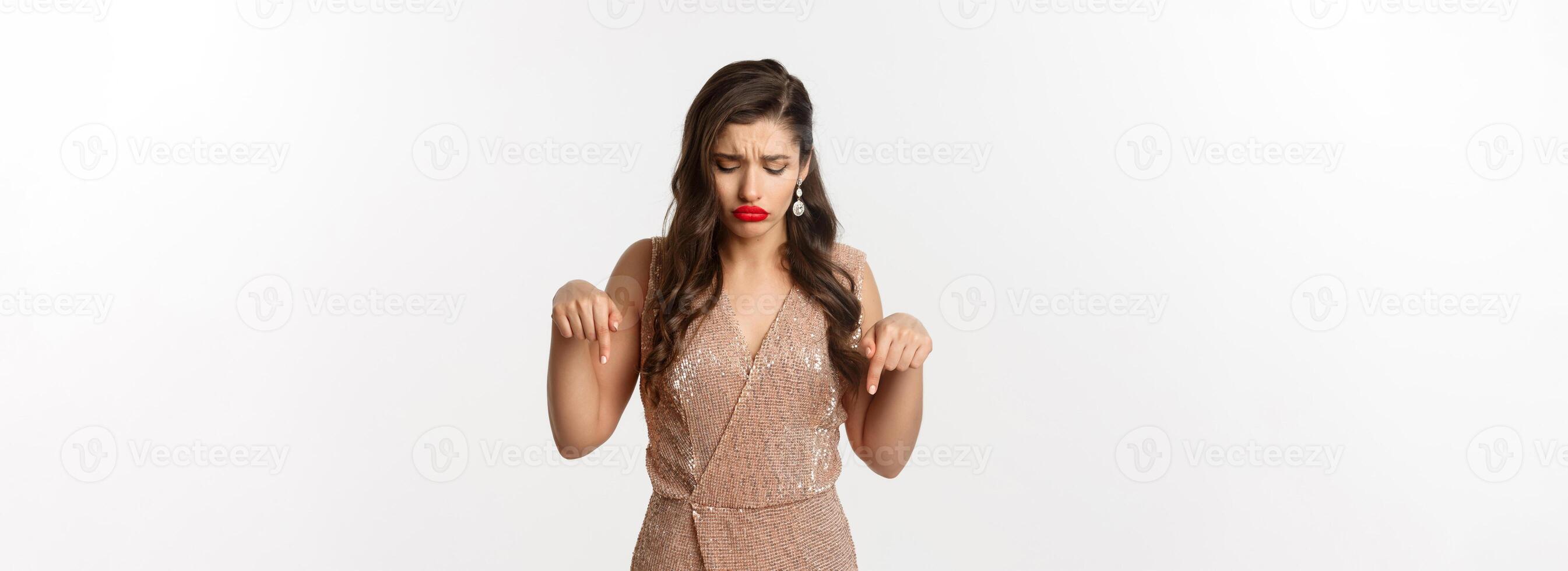 Sad beautiful girl pointing fingers down and sulking, wearing dress for christmas party, looking disappointed, standing over white background photo