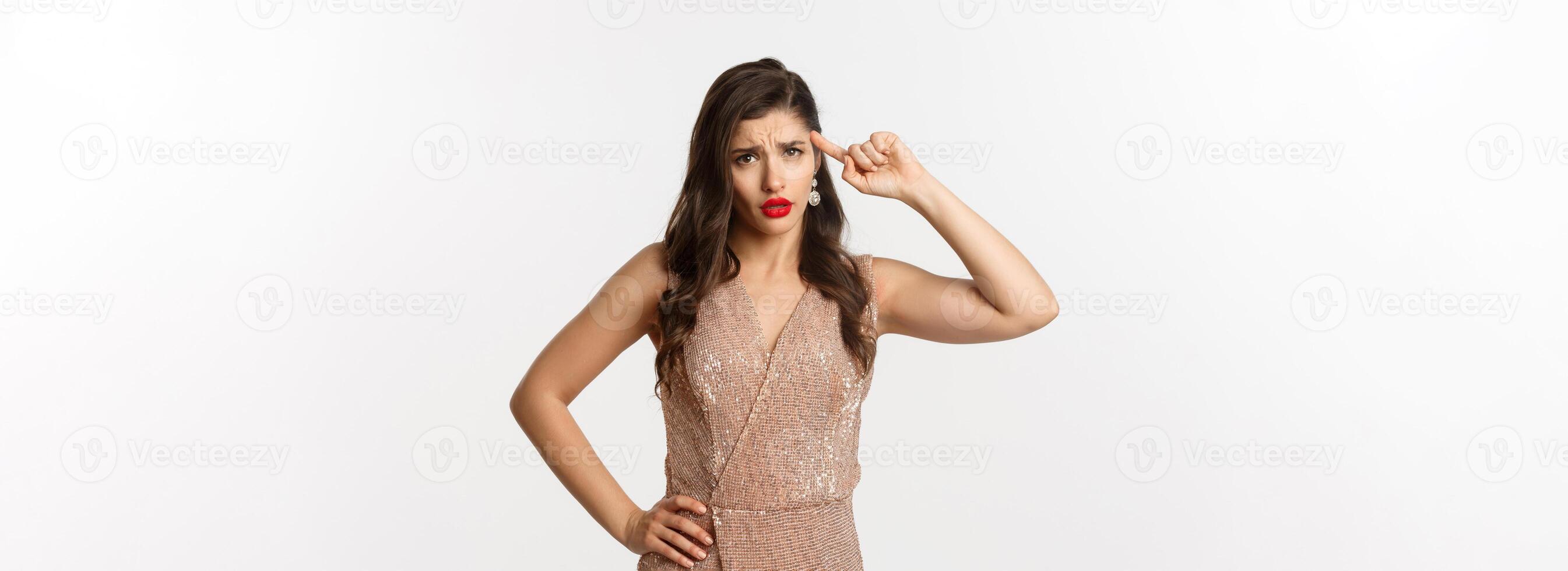 Concept of casino, celebration and party. Woman looking with disdain and roll finger on head, scolding someone, are you stupid gesture, standing in dress over white background photo