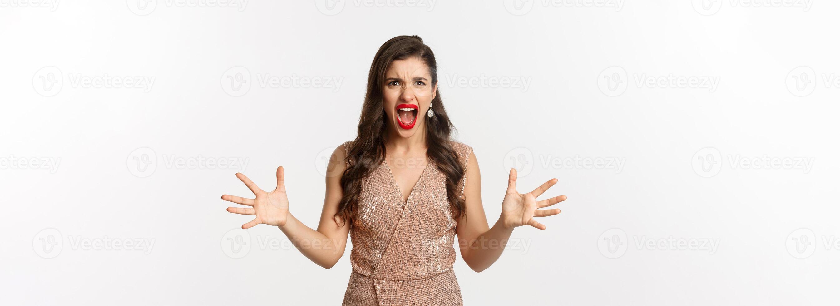 Celebration and party concept. Angry young woman in glamour dress shouting at you, looking mad and outraged, shaking hands aggressive, having an argument, white background photo