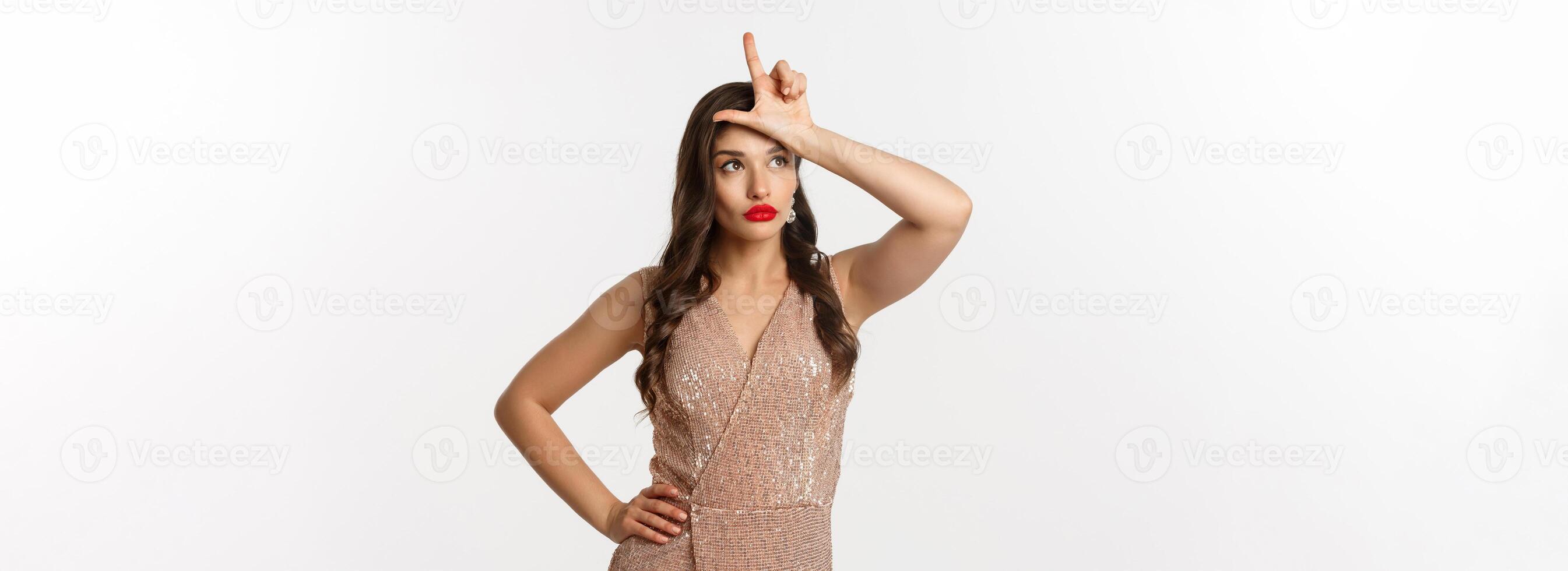 Celebration and party concept. Attractive young woman with red lips, glamour dress, showing loser sign on forehead and looking away ignorant, standing over white background photo