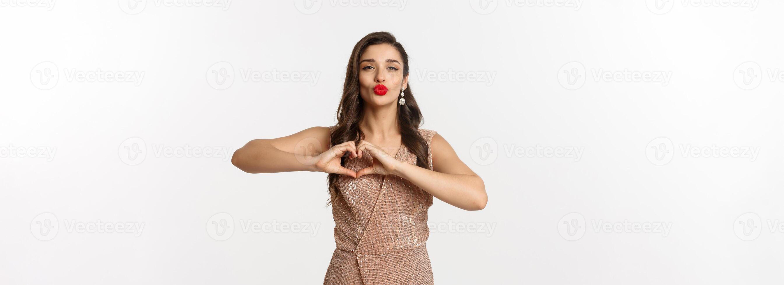Celebration and party concept. Beautiful young woman in stylish dress, red lips, showing heart sign and waiting for kiss, standing over white background photo