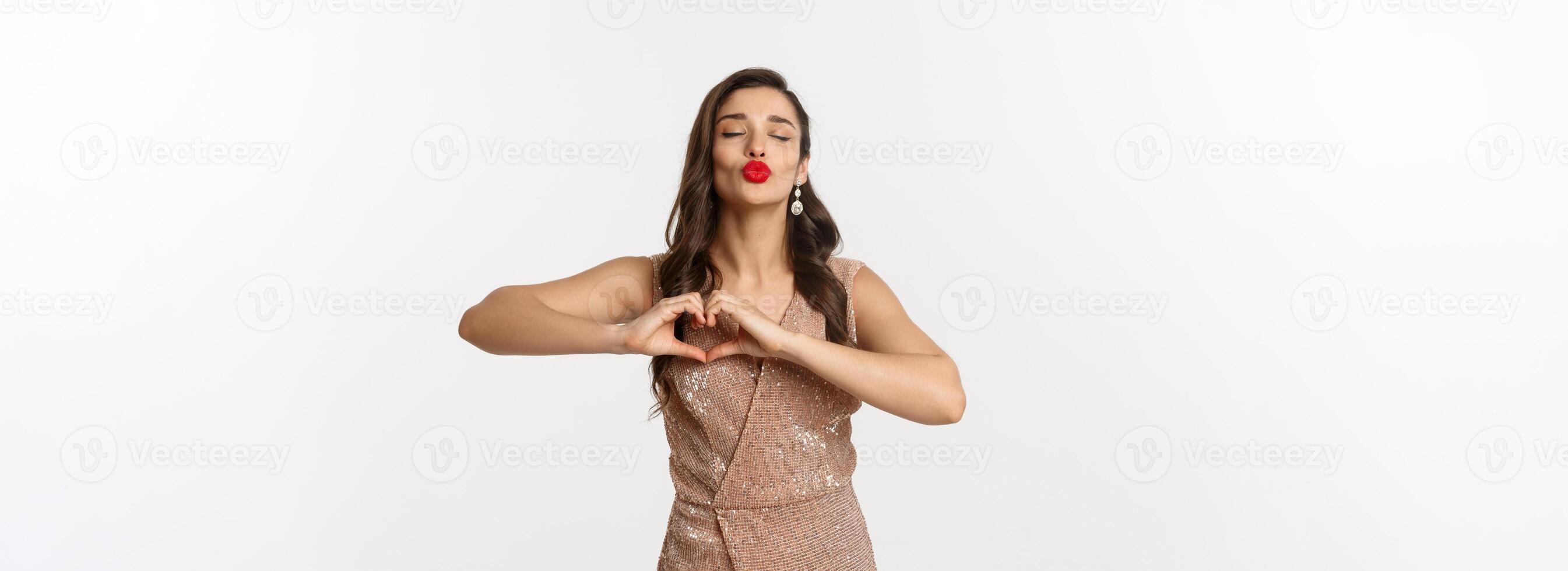 Celebration and party concept. Beautiful young woman in stylish dress, red lips, showing heart sign and waiting for kiss, standing over white background photo