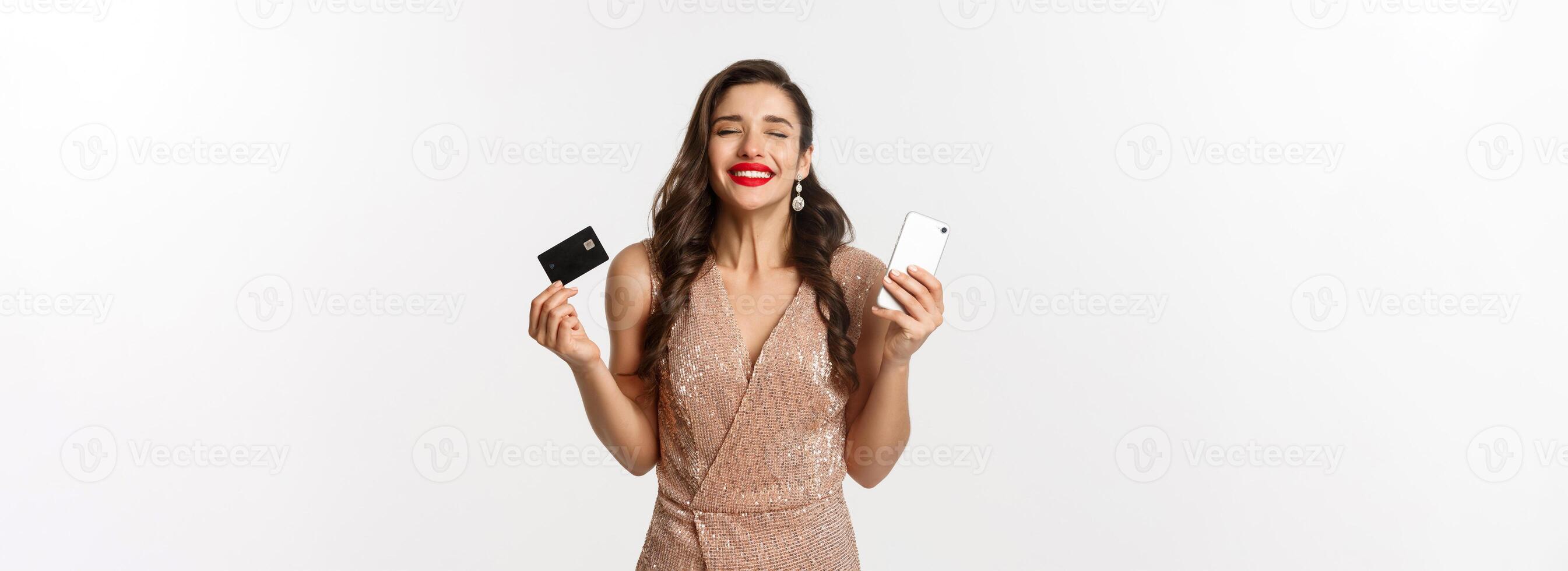 Online shopping and holidays concept. Satisfied and happy woman in elegant dress smiling, using credit card and mobile phone, standing over white background photo
