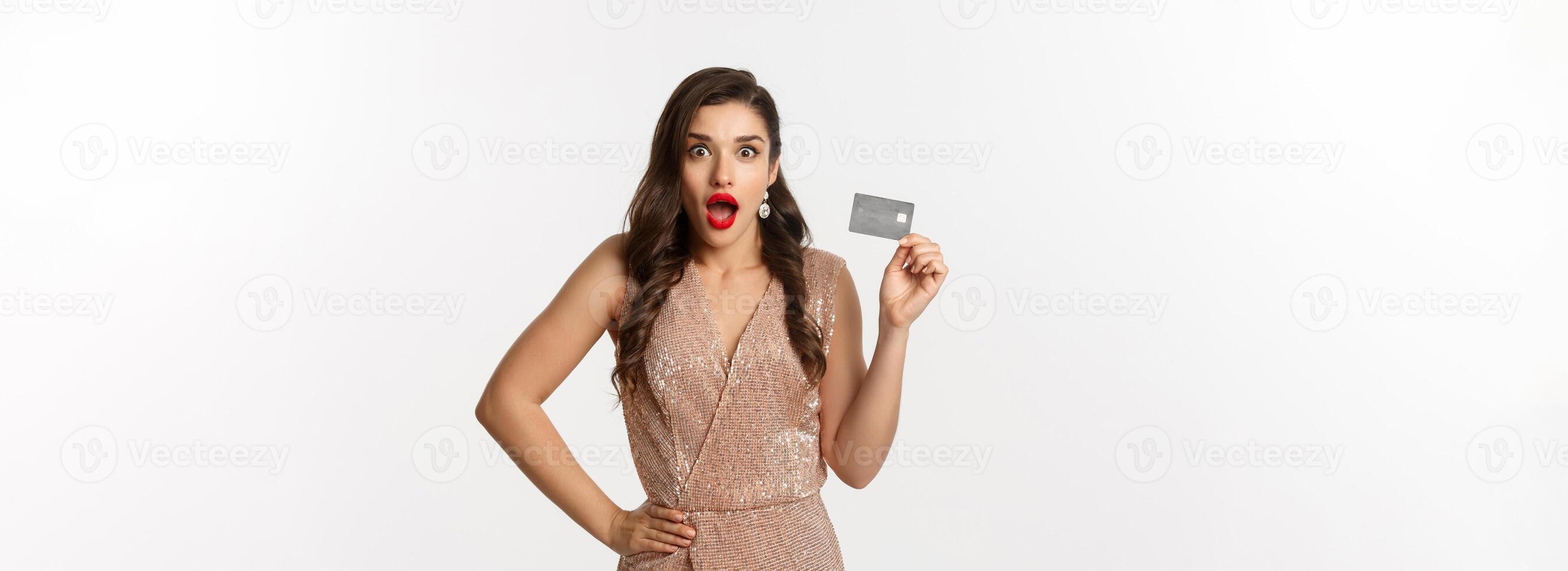 Holidays. christmas party and shopping concept. Amazed attractive woman with red lips, luxury dress, showing credit card and staring at camera, standing over white background photo