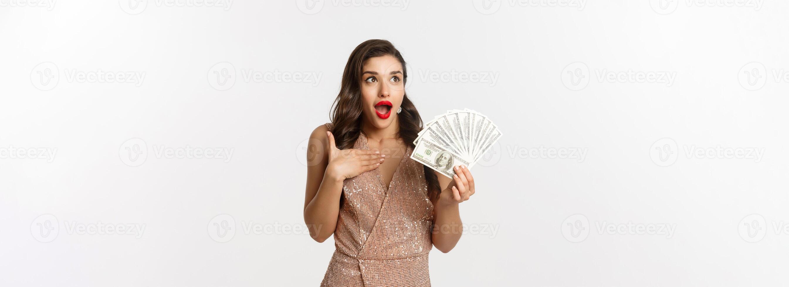 Shopping concept. Image of surprised and excited woman holding money, staring at upper left corner amazed, standing with dollars in glamour dress, white background photo