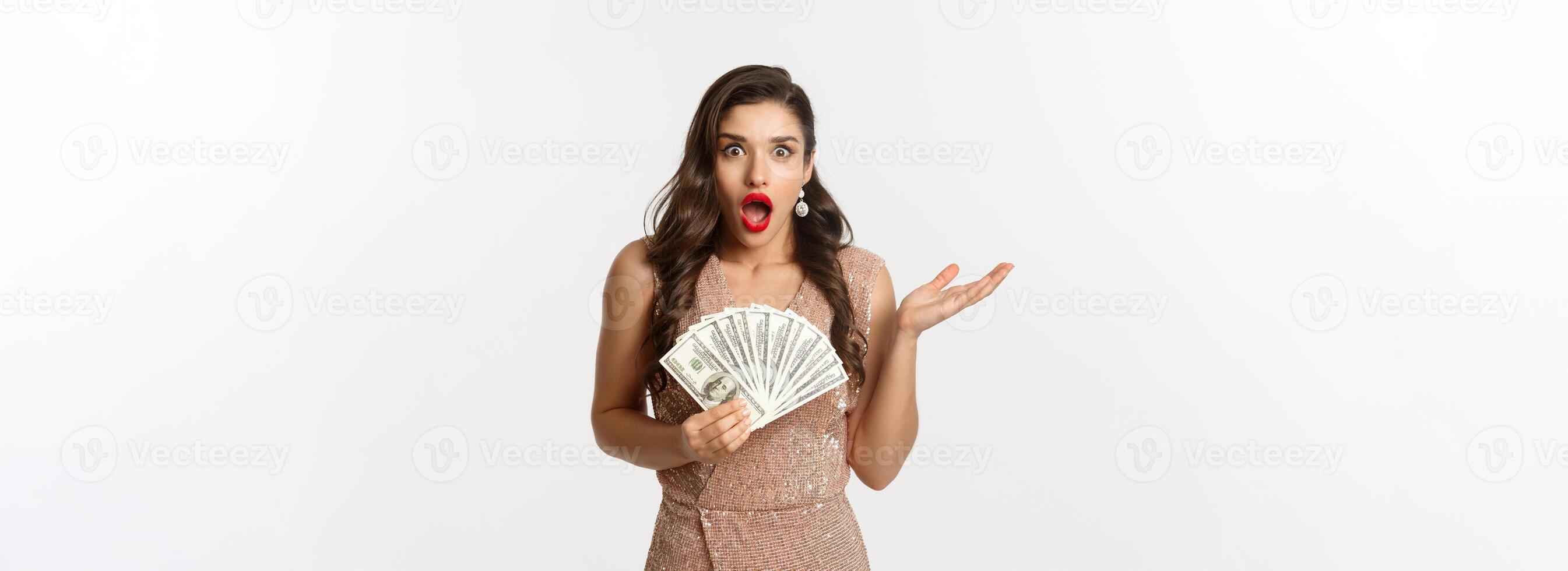 Shopping concept. Image of beautiful woman in glamour dress looking shocked, holding dollars and staring at camera, standing over white background photo