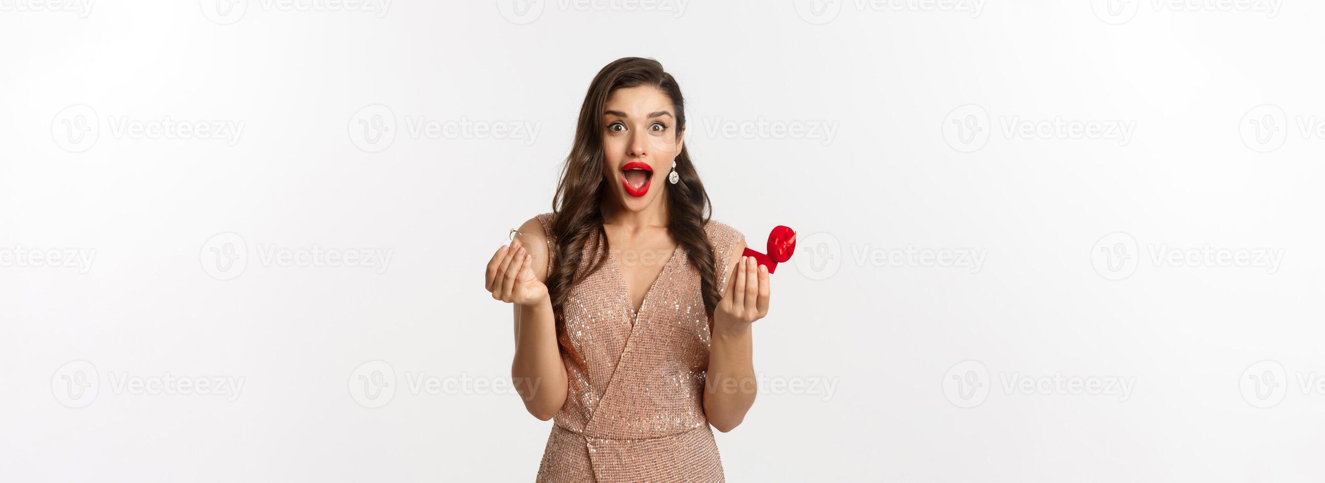 Excited woman holding engagement ring and red box, looking surprised and amazed, receive marriage proposal on date, wearing elegant dress, standing over white background photo