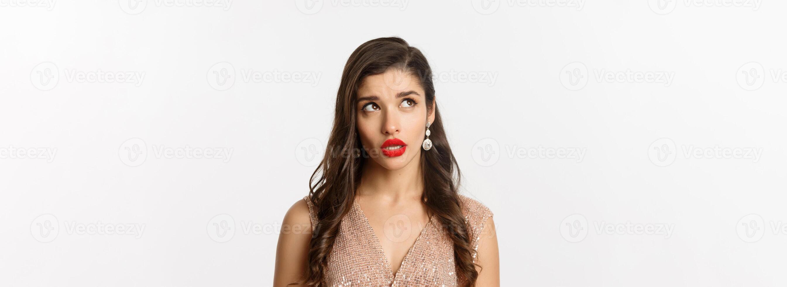 Concept of New Year celebration and winter holidays. Close-up of elegant woman with red lips and dress, looking at upper left corner and thinking, standing over white background photo