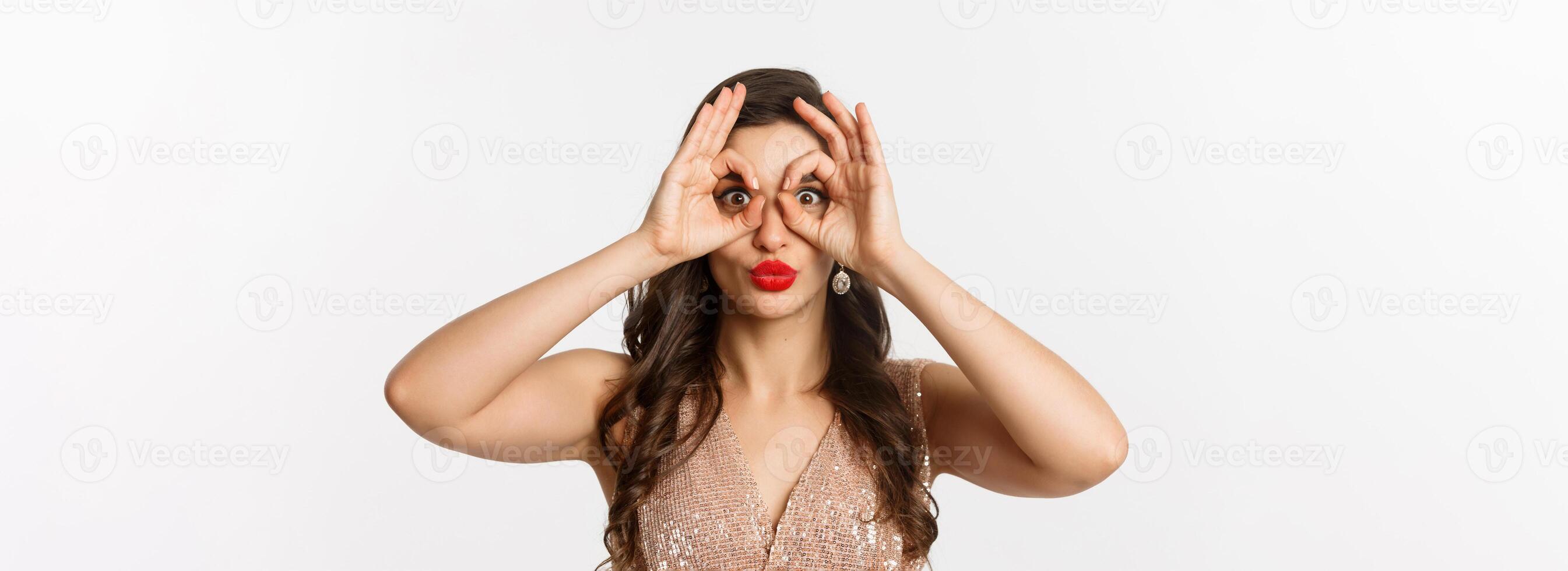 Concept of New Year celebration and winter holidays. Close-up of beautiful brunette woman in dress, red lips, making hand binoculars and staring at camera, white background photo