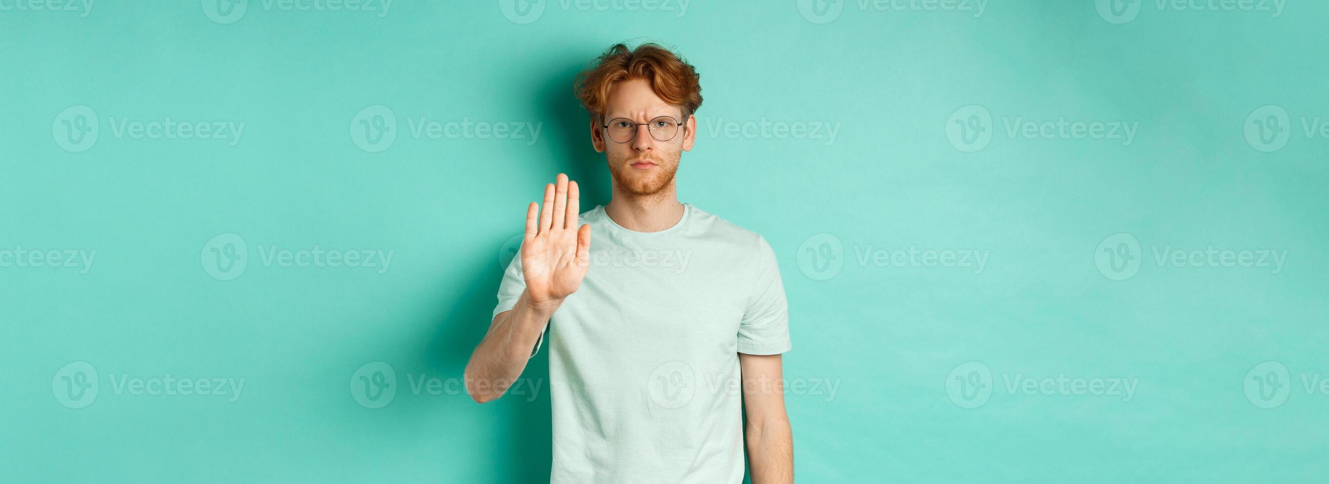 Angry and serious young man with red hair, wearing glasses, showing stop gesture, telling no, disapprove and prohibit something bad, standing over turquoise background photo