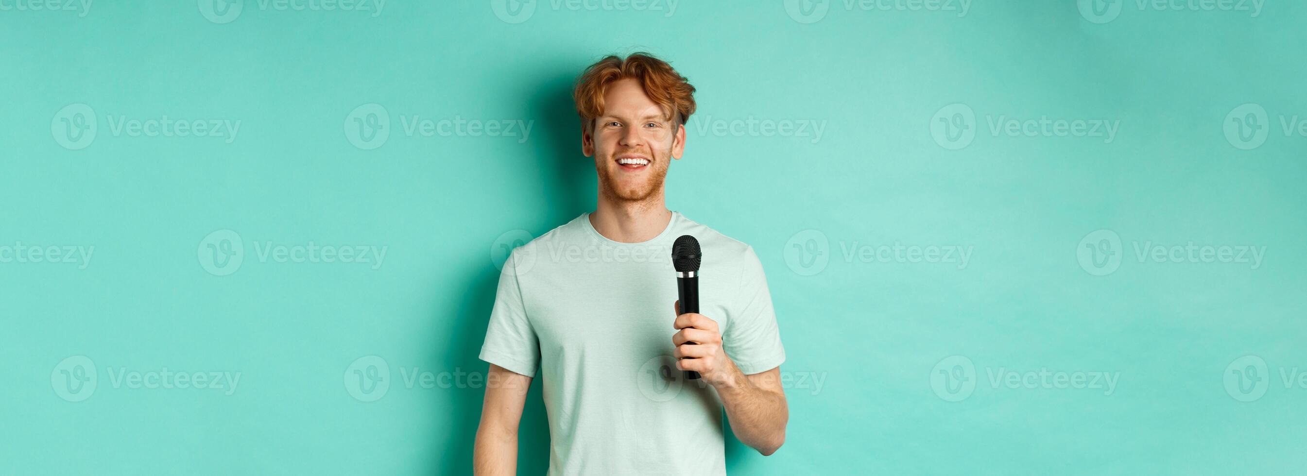 Young redhead man with beard, wearing t-shirt, holding microphone and making speech, singing karaoke, standing over mint background photo