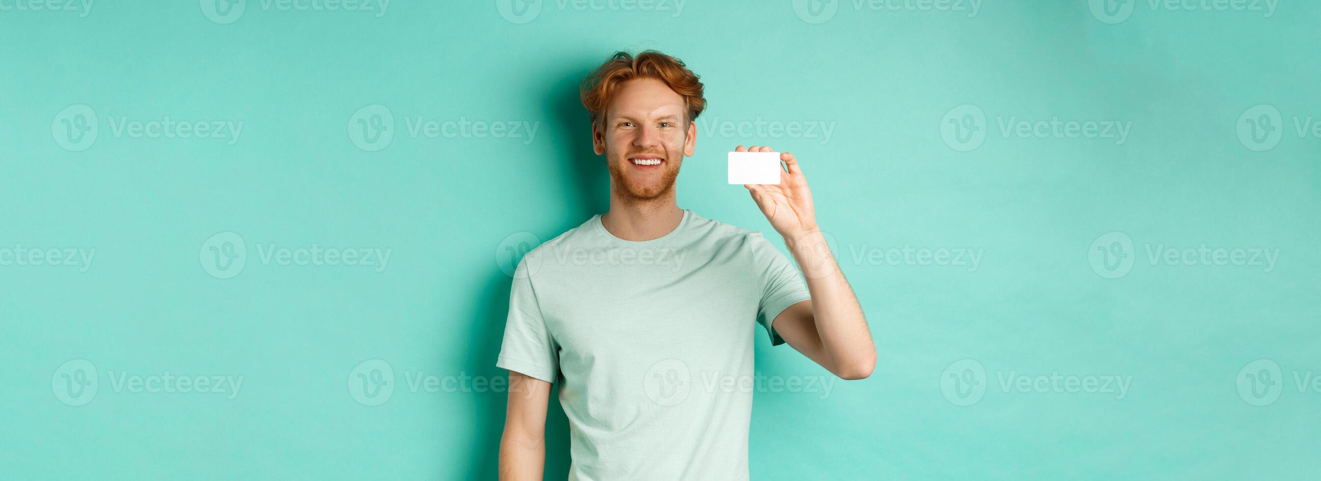 Shopping concept. Handsome redhead man in t-shirt showing plastic credit card and smiling, standing over turquoise background photo