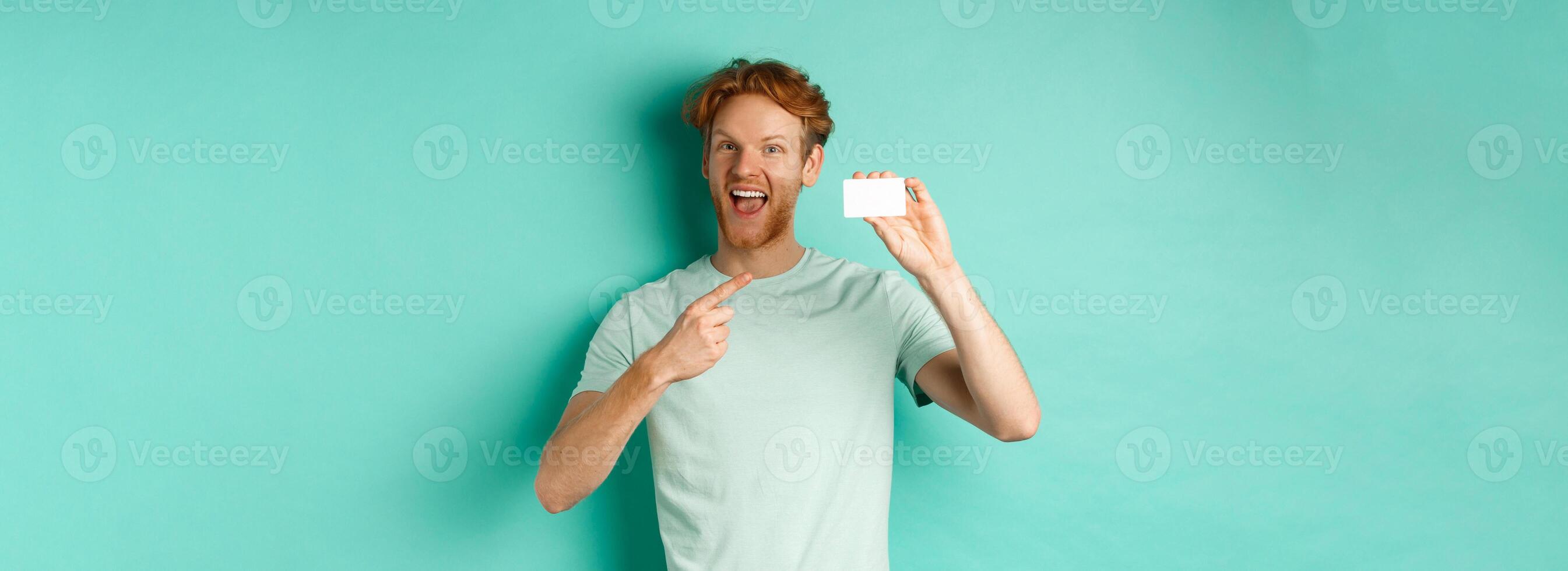Shopping concept. Handsome redhead man in t-shirt showing plastic credit card and smiling, standing over turquoise background photo