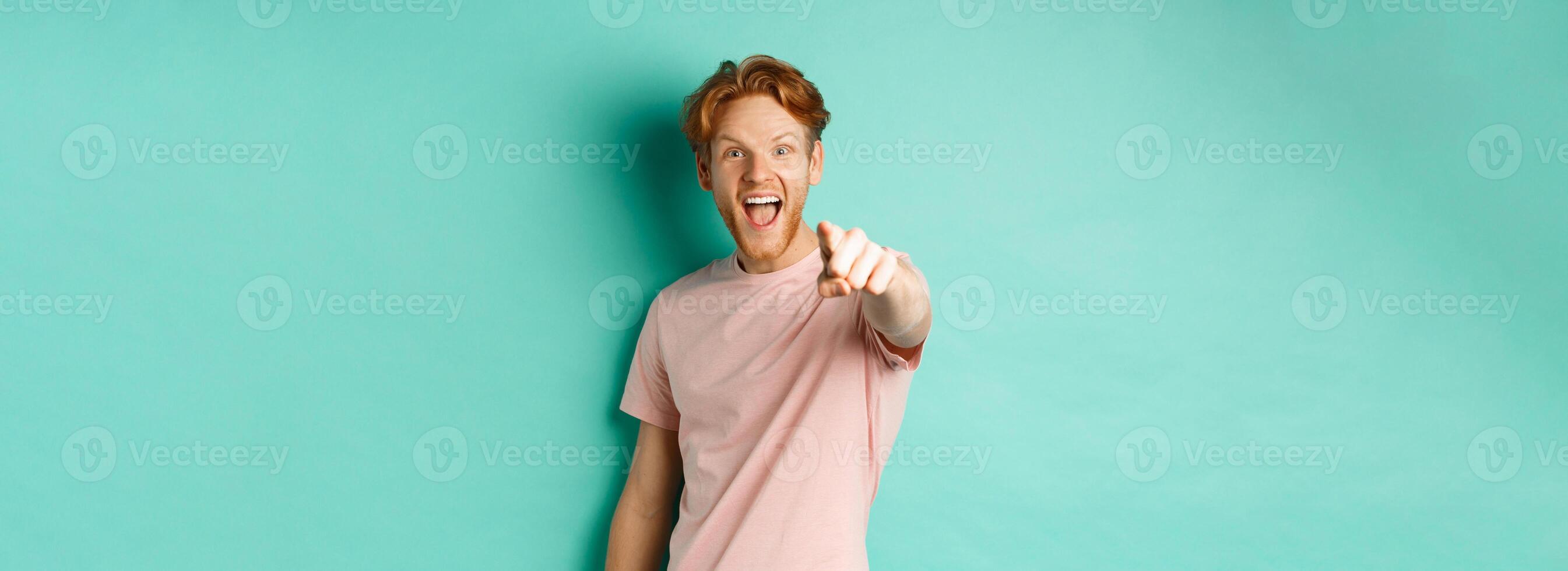 Excited young man with red hair checking out something cool, pointing finger at camera and smiling fascinated, standing over turquoise background photo