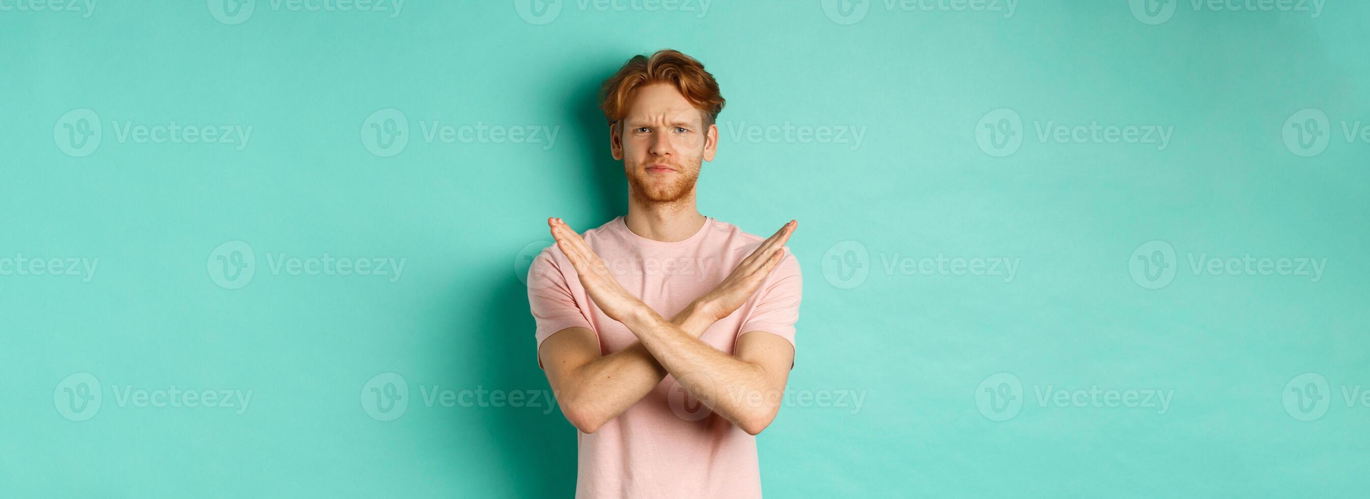 Skeptical redhead man in t-shirt saying no, cross arms on chest and frowning disappointed, prohibit something bad, standing over turquoise background photo