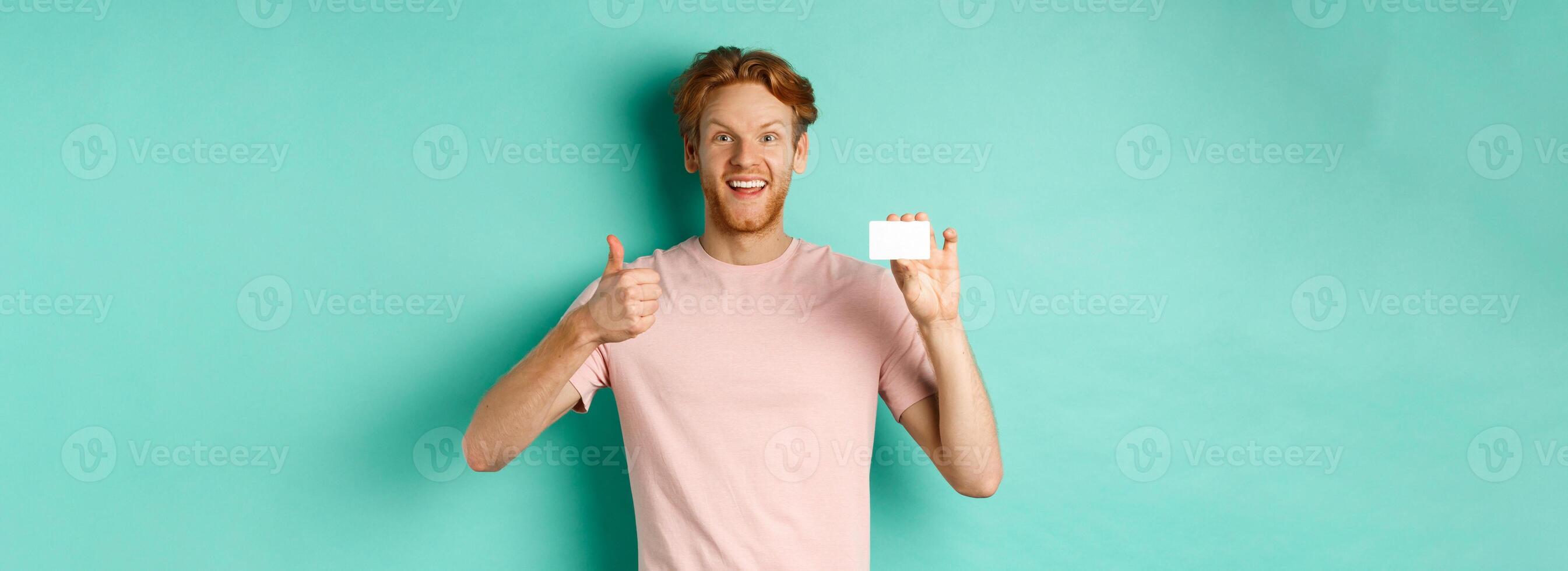Cheerful male bank client in t-shirt showing thumb up and plastic credit card, smiling satisfied at camera, standing over turquoise background photo