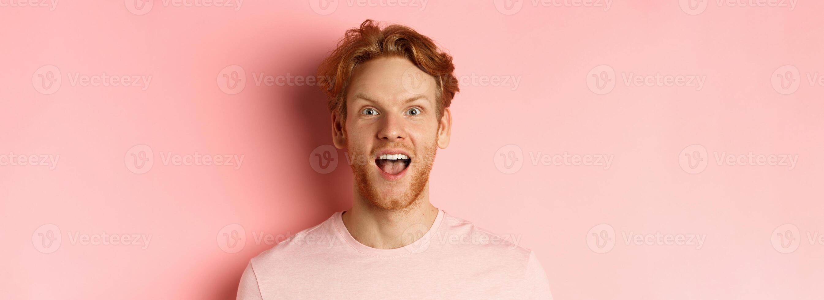 Headshot of amazed redhead male model in t-shirt open mouth, looking fascinated and happy at camera, standing over pink background photo