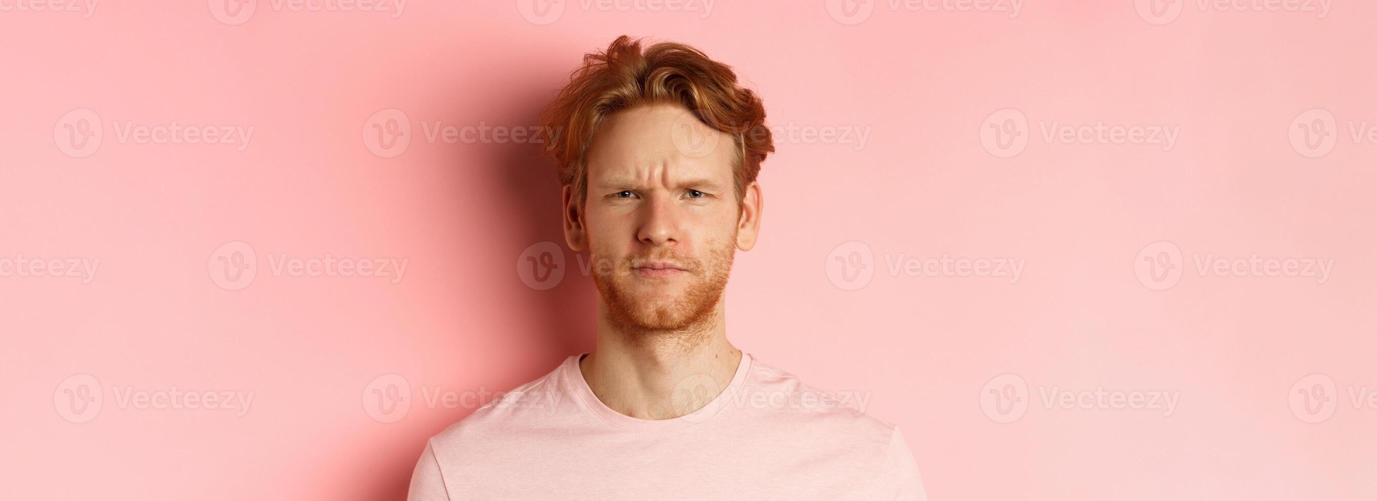 Close up of suspicious redhead guy frowning, staring at camera doubtful, express disapproval, standing over pink background photo