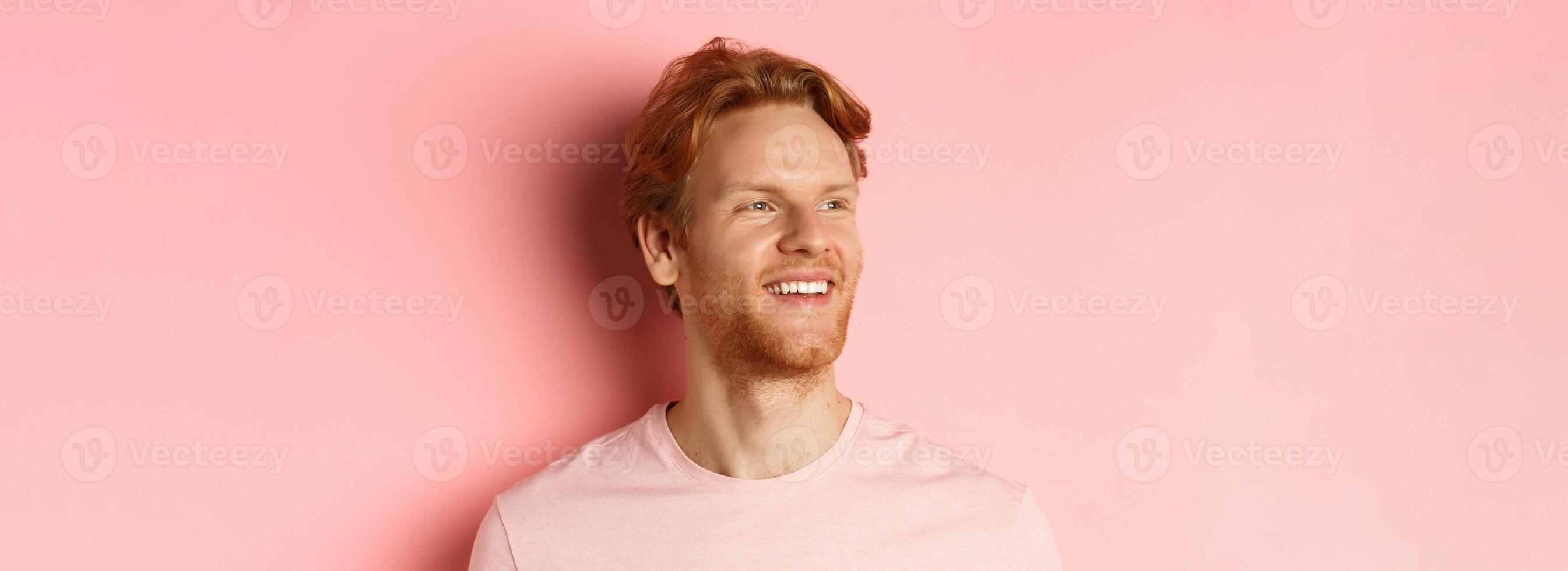 Close up of handsome young man with beard and red hair, looking left and smiling delighted, standing confident over pink background photo