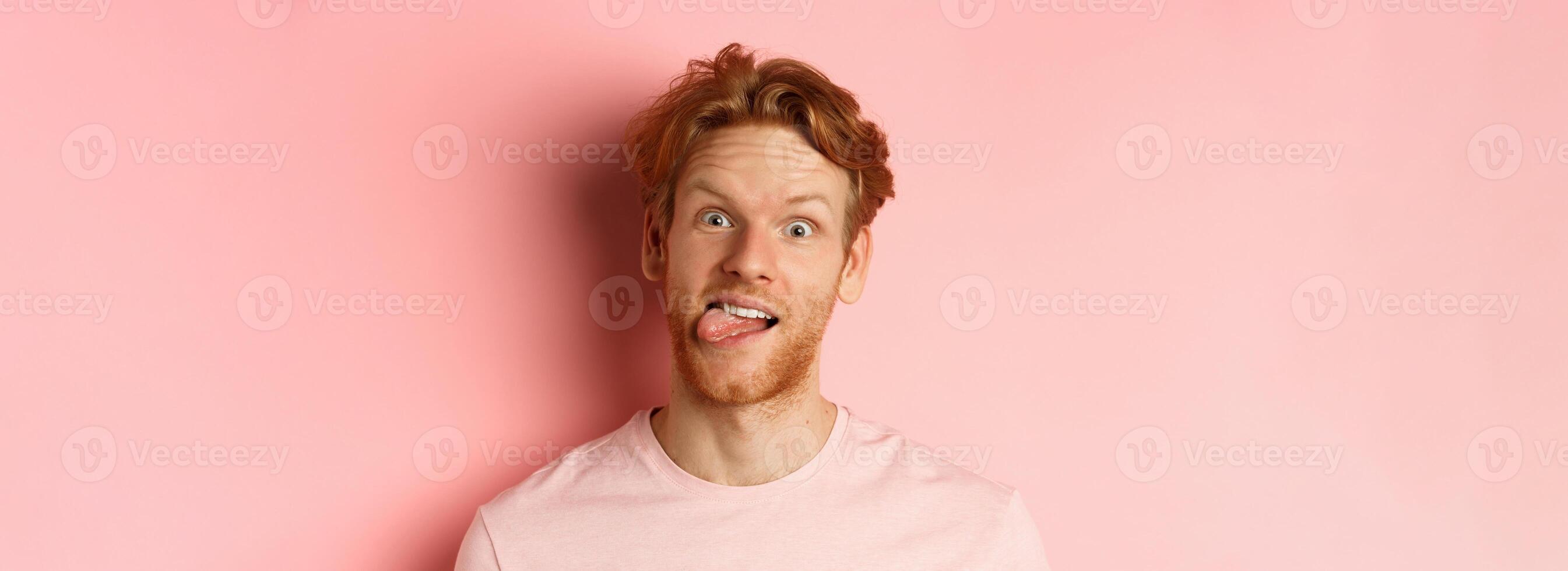 Headshot of funny redhead guy showing tongue, making silly faces at camera, standing joyful against pink background photo