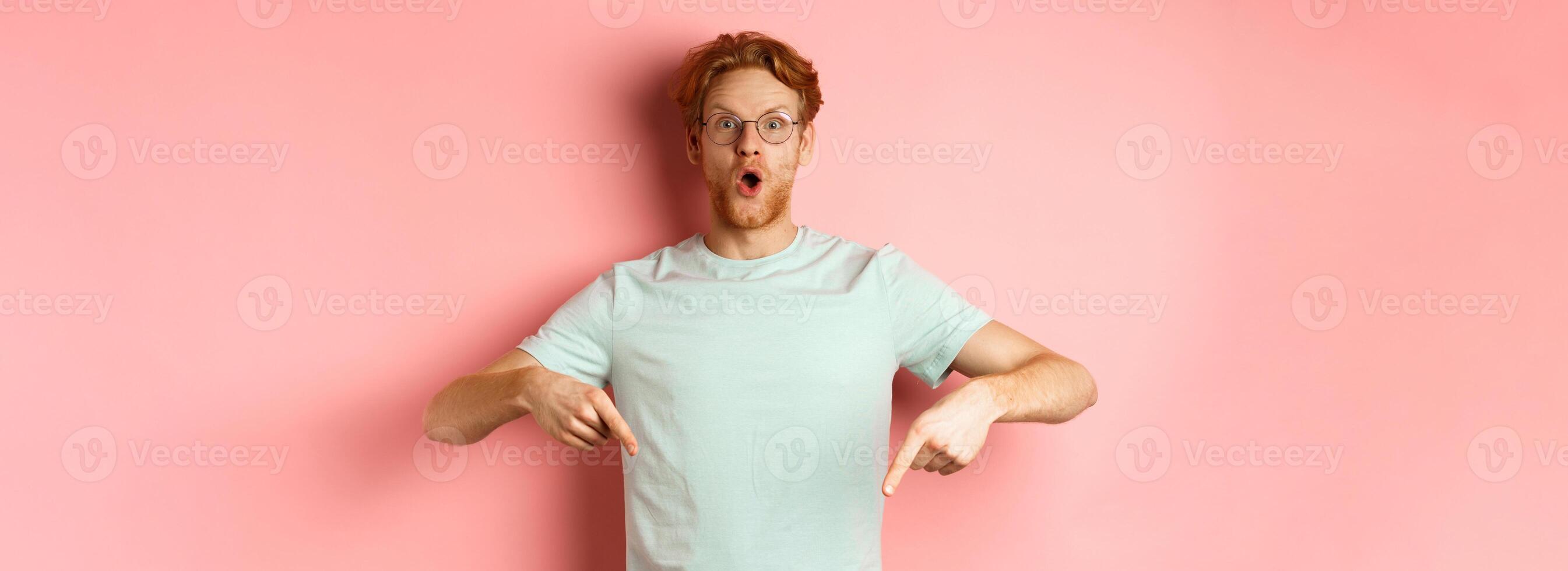Image of amazed young man with red hair and beard, wearing glasses and t-shirt, pointing fingers down and staring excited at camera, pink background photo