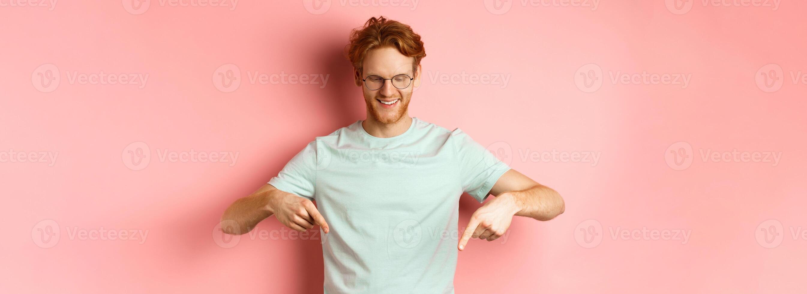 Image of handsome caucasian man with red messy hair and glasses, pointing fingers and looking down at promo offer, smiling pleased, standing over pink background photo