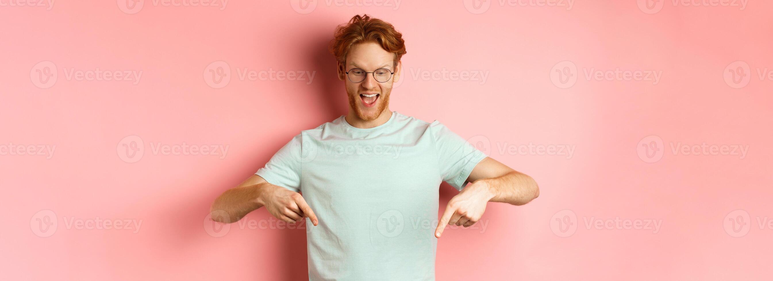 Handsome caucasian man with red messy hair and glasses, pointing fingers and looking down at promo offer, smiling pleased, standing over pink background photo