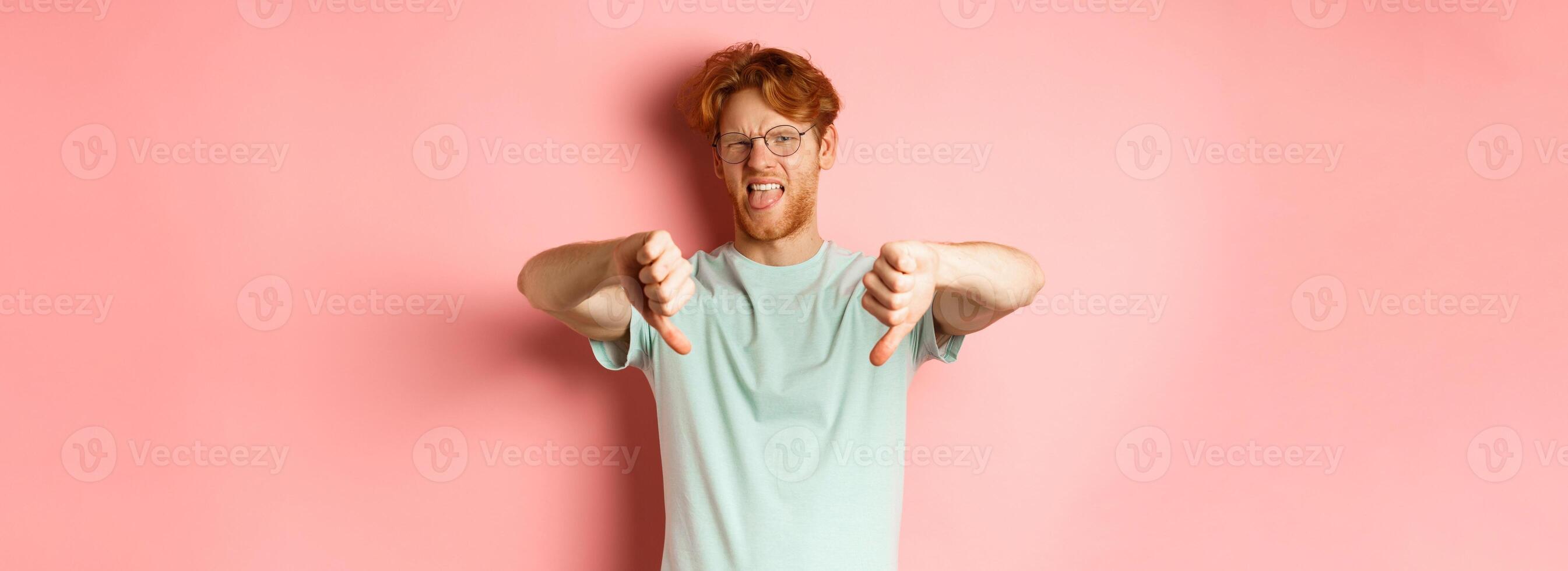 Disgusted redhead man in t-shirt and glasses showing thumbs down and tongue, grimacing from aversion, standing over pink background photo
