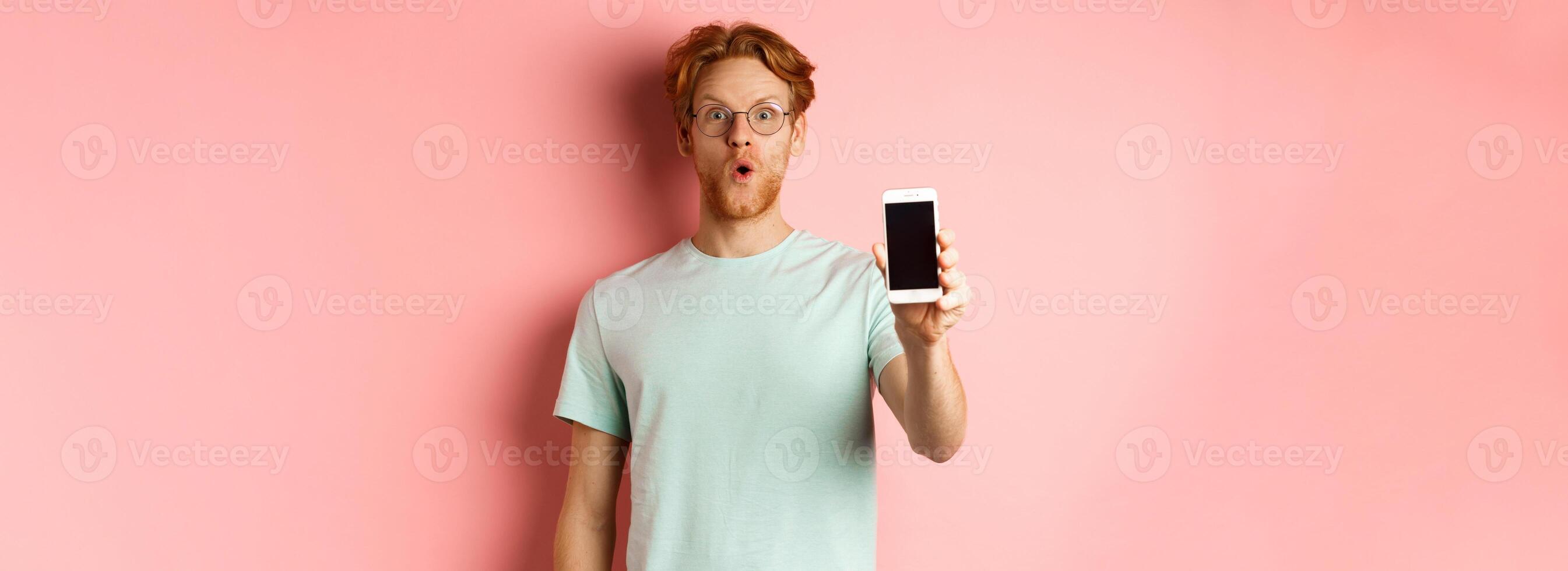 Image of handsome guy with red hair, wearing glasses and t-shirt, saying wow and showing smartphone screen, standing against pink background photo