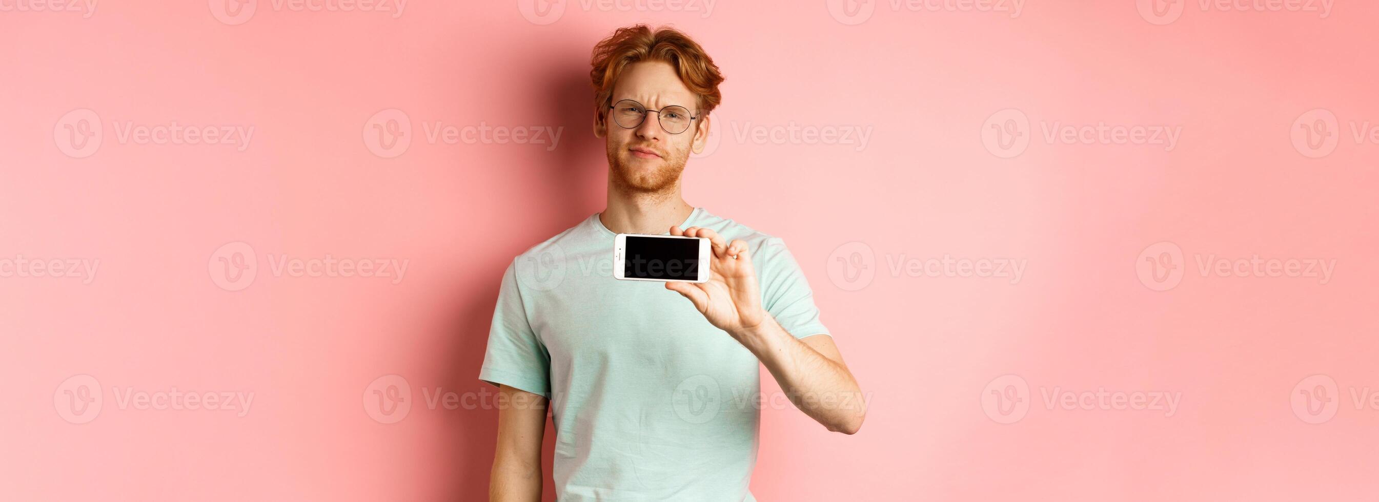 Skeptical redhead guy in glasses showing smartphone screen horizontally, smirk and frowning disappointed, standing over pink background photo