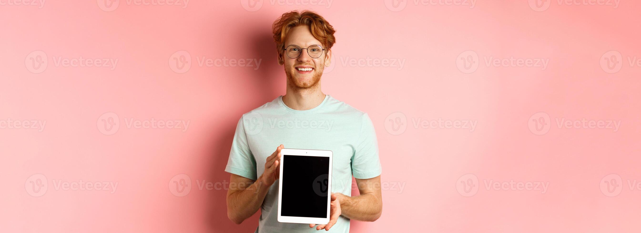 Funny redhead guy in glasses demonstrate digital tablet screen, smiling at camera, standing over pink background photo