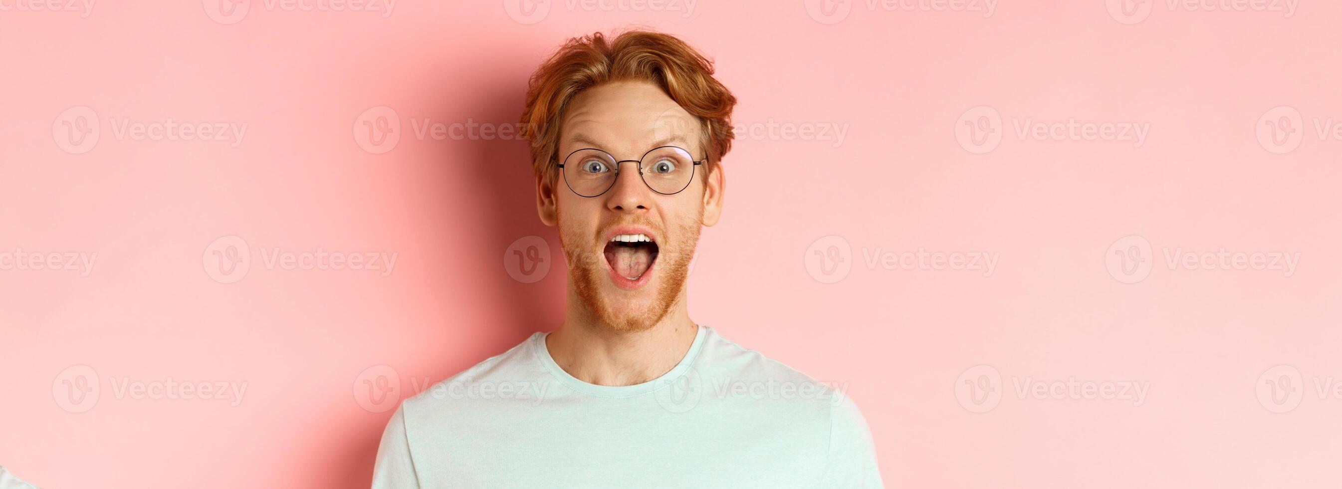 Close up of surprised redhead man raising eyebrows and scream with joy, checking out cool promo, looking amazed at camera, standing over pink background photo