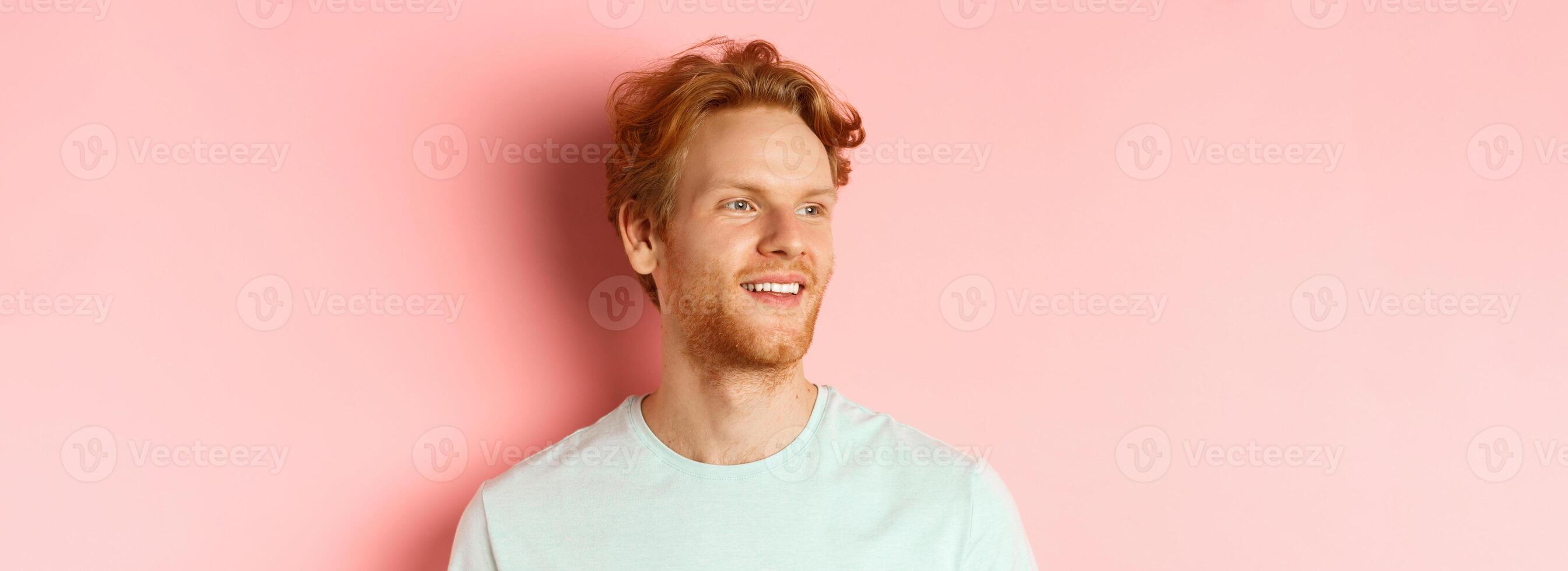 Portrait of attractive caucasian guy with red messy hair and beard, turn head and looking left with pleased smile, standing over pink background photo
