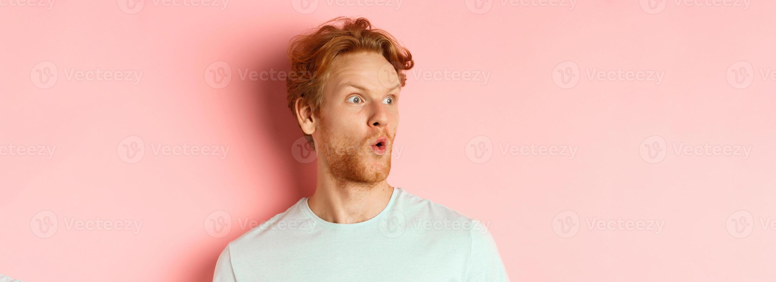 Headshot portrait of surprised redhead man with beard, looking left and saying wow, raising eyebrows amazed, standing over pink background photo