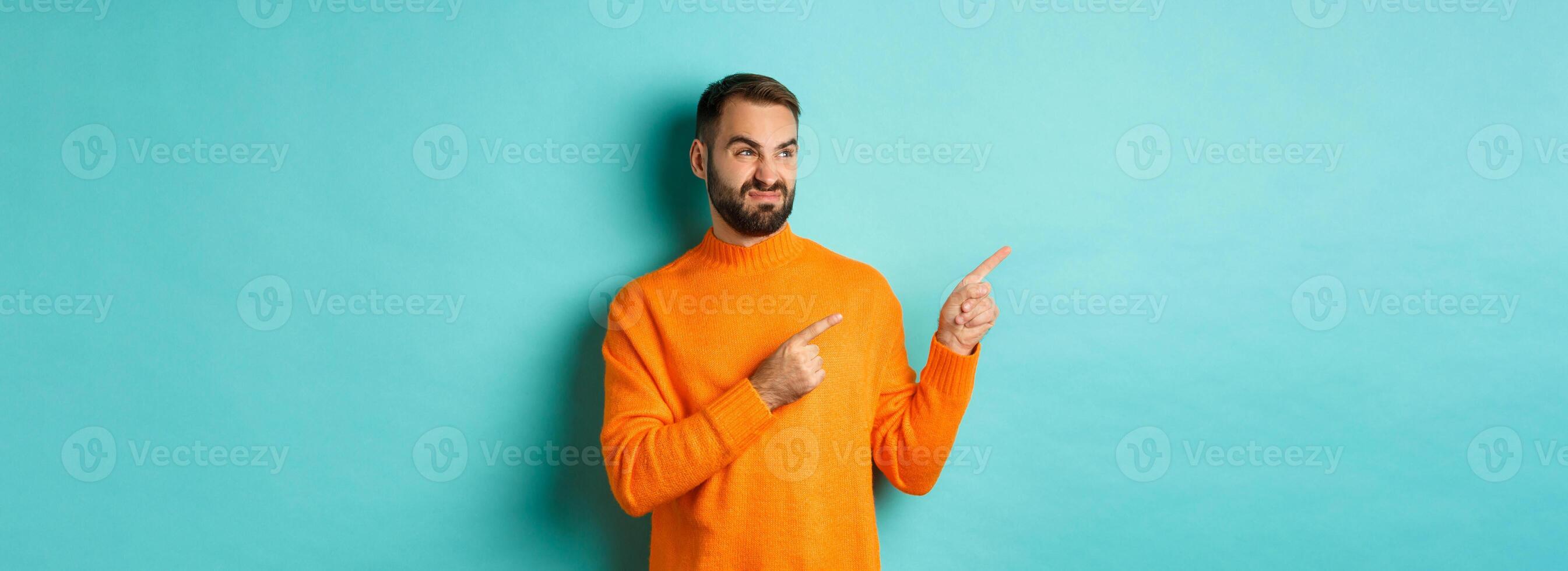 Displeased and skeptical guy grimacing, showing bad product, pointing fingers left with disappointment, standing turquoise background photo